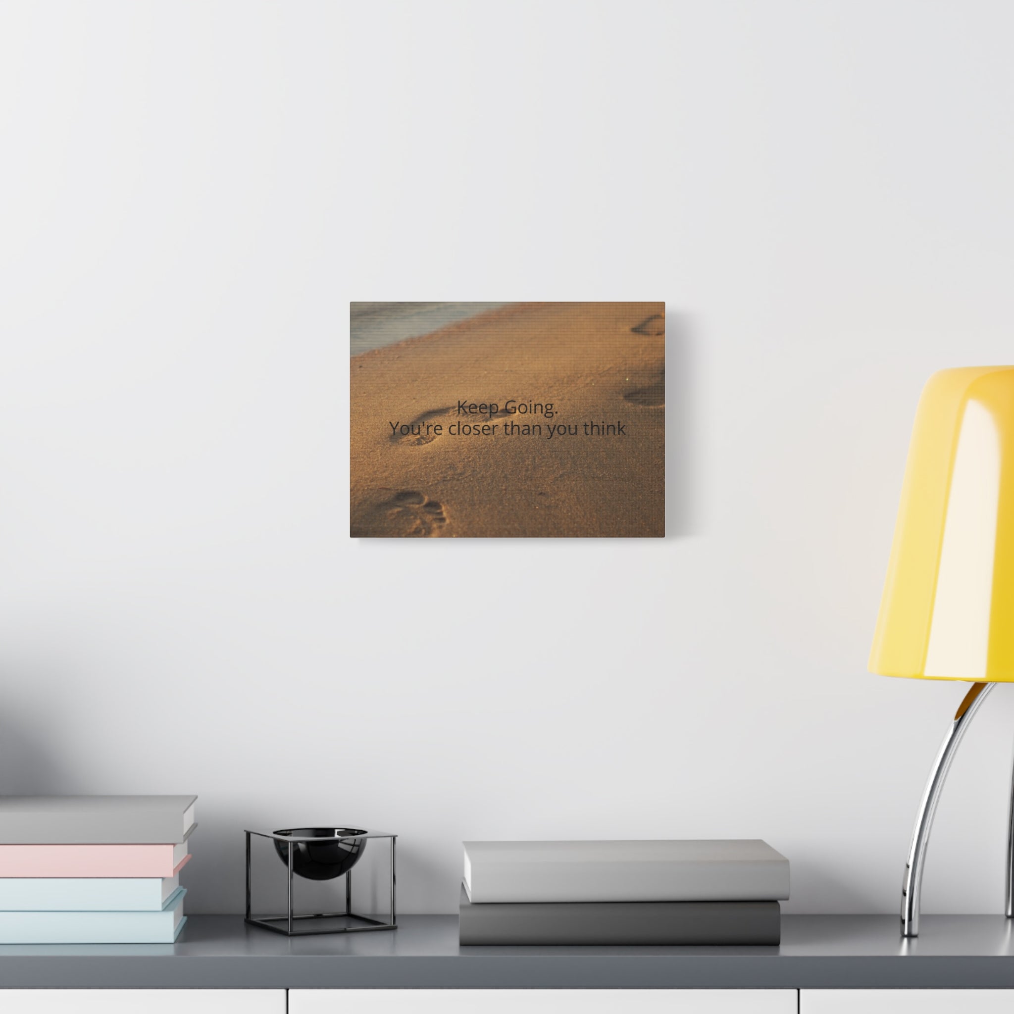 Wall art with motivational quote on a white wall above a desk with books and a lamp.
