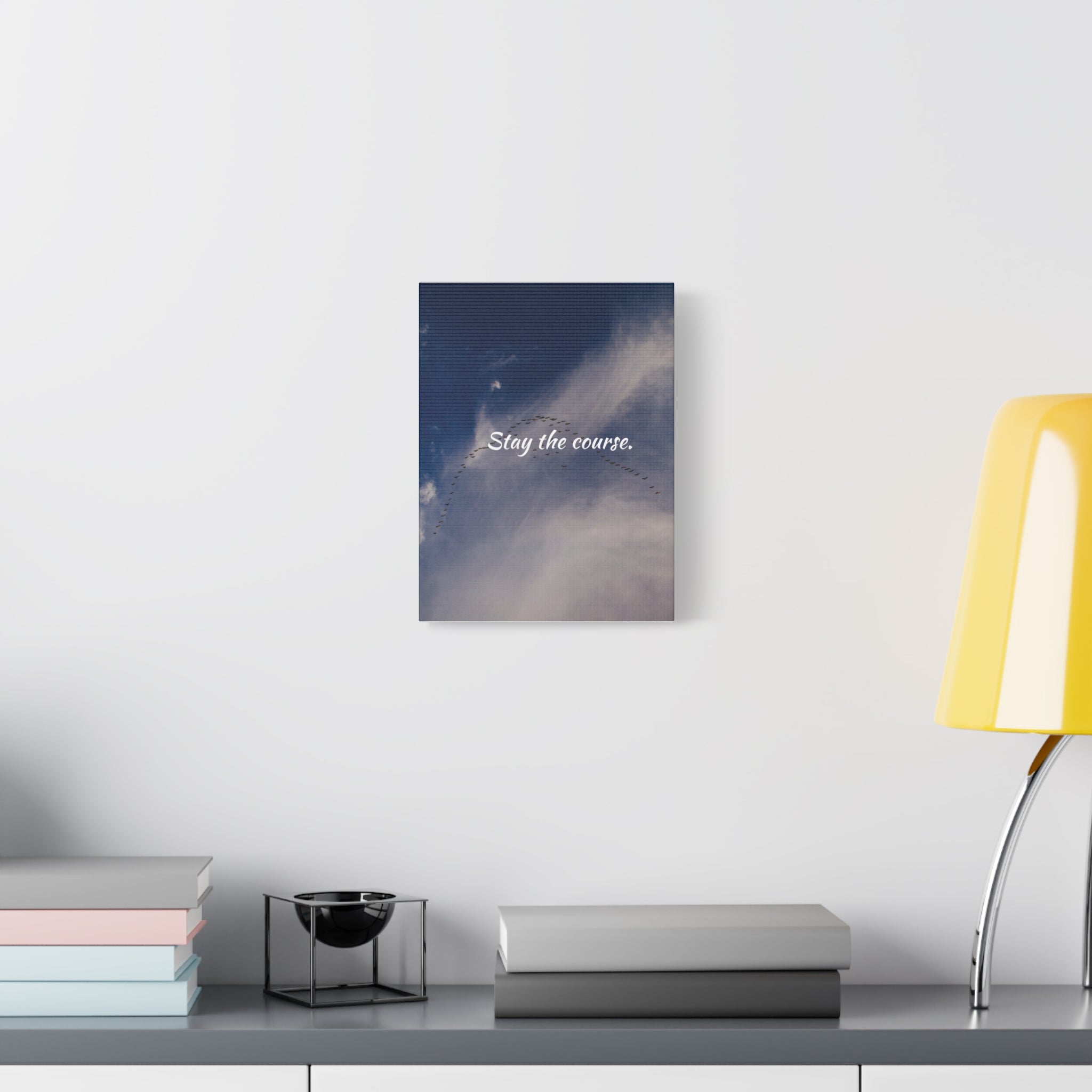 Canvas print with 'Stay the course.' text on a wall above a desk with books and a lamp.