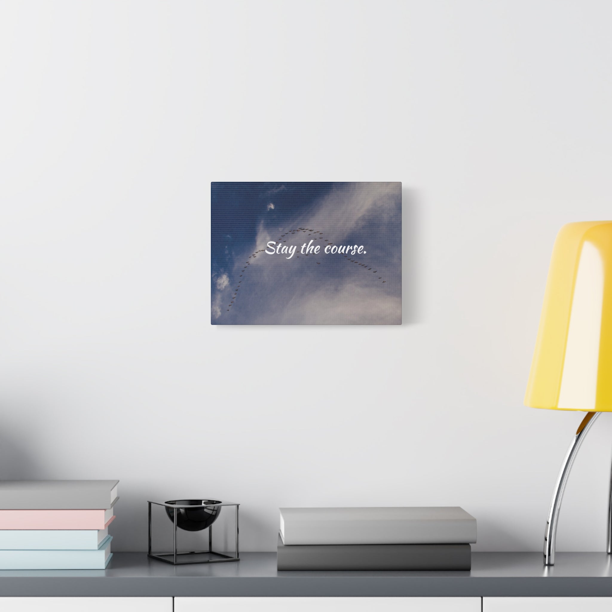 Wall art with 'Stay the course.' text on a celestial background above a desk with books and a lamp.