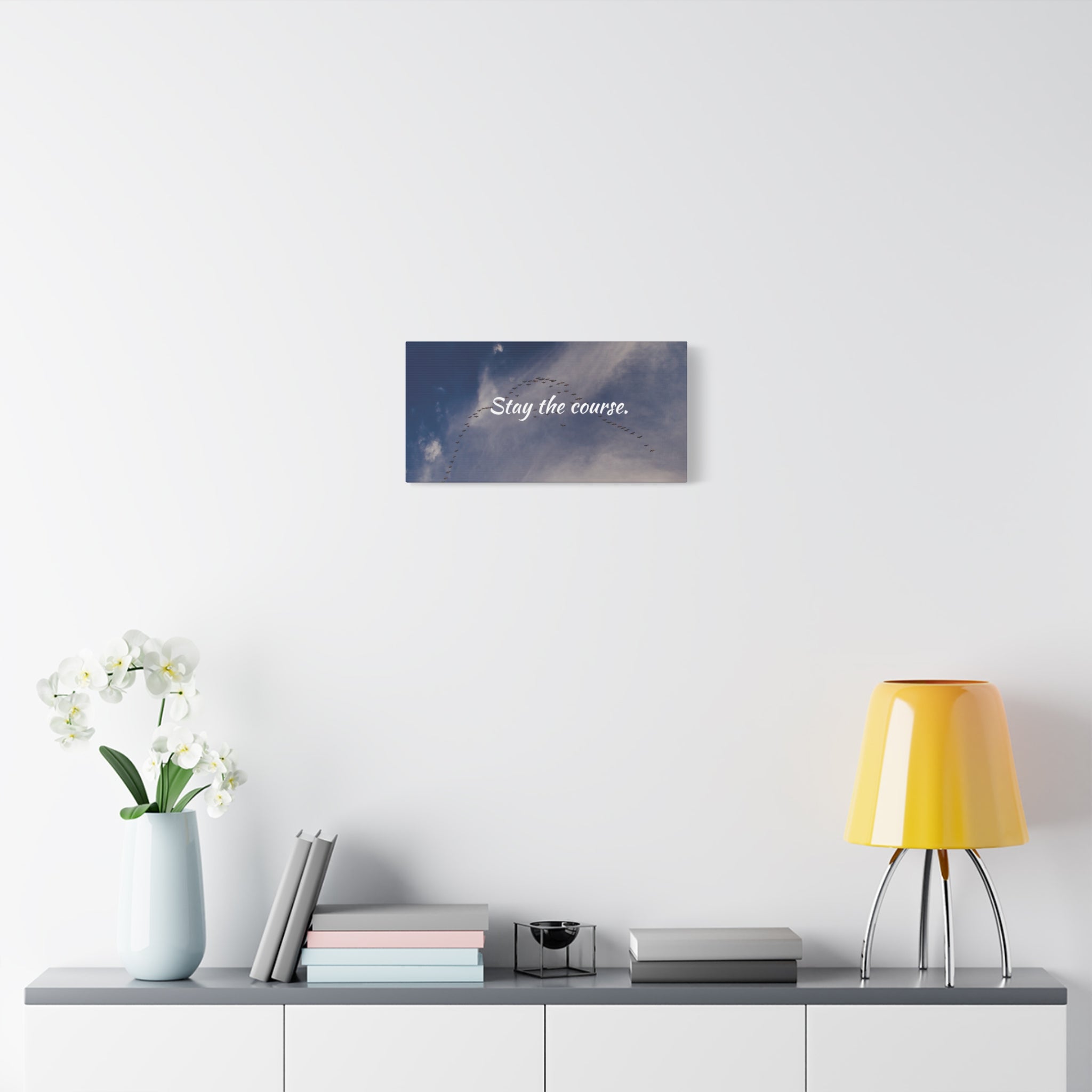 Decorative wall art with motivational quote 'Stay the course' on a white wall above a shelf with books and a lamp.