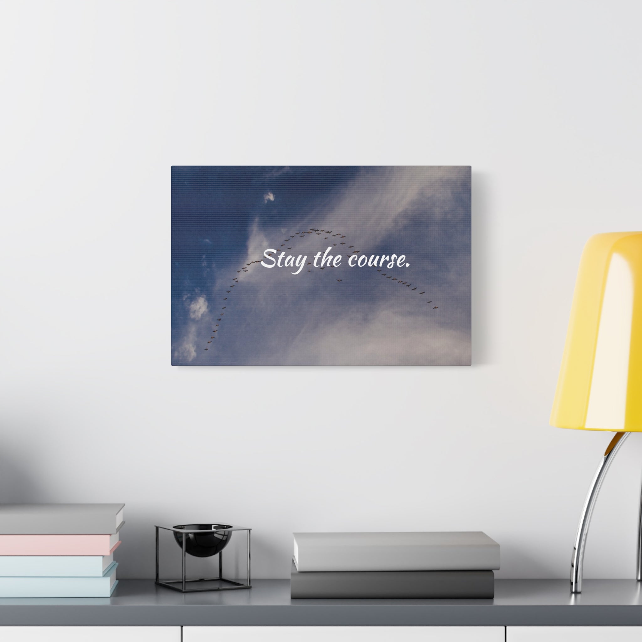 Canvas print with 'Stay the course.' text on a wall above a desk with books and a lamp.