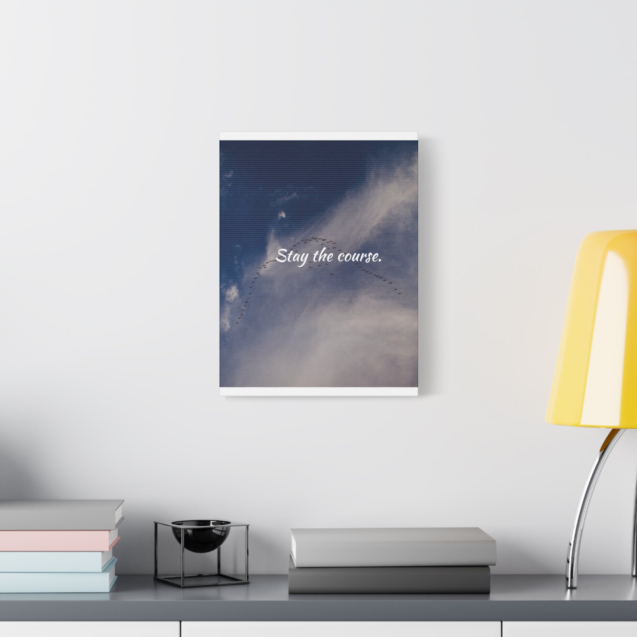 Framed artwork with 'Stay the course.' text on a wall above a desk with books and a lamp.