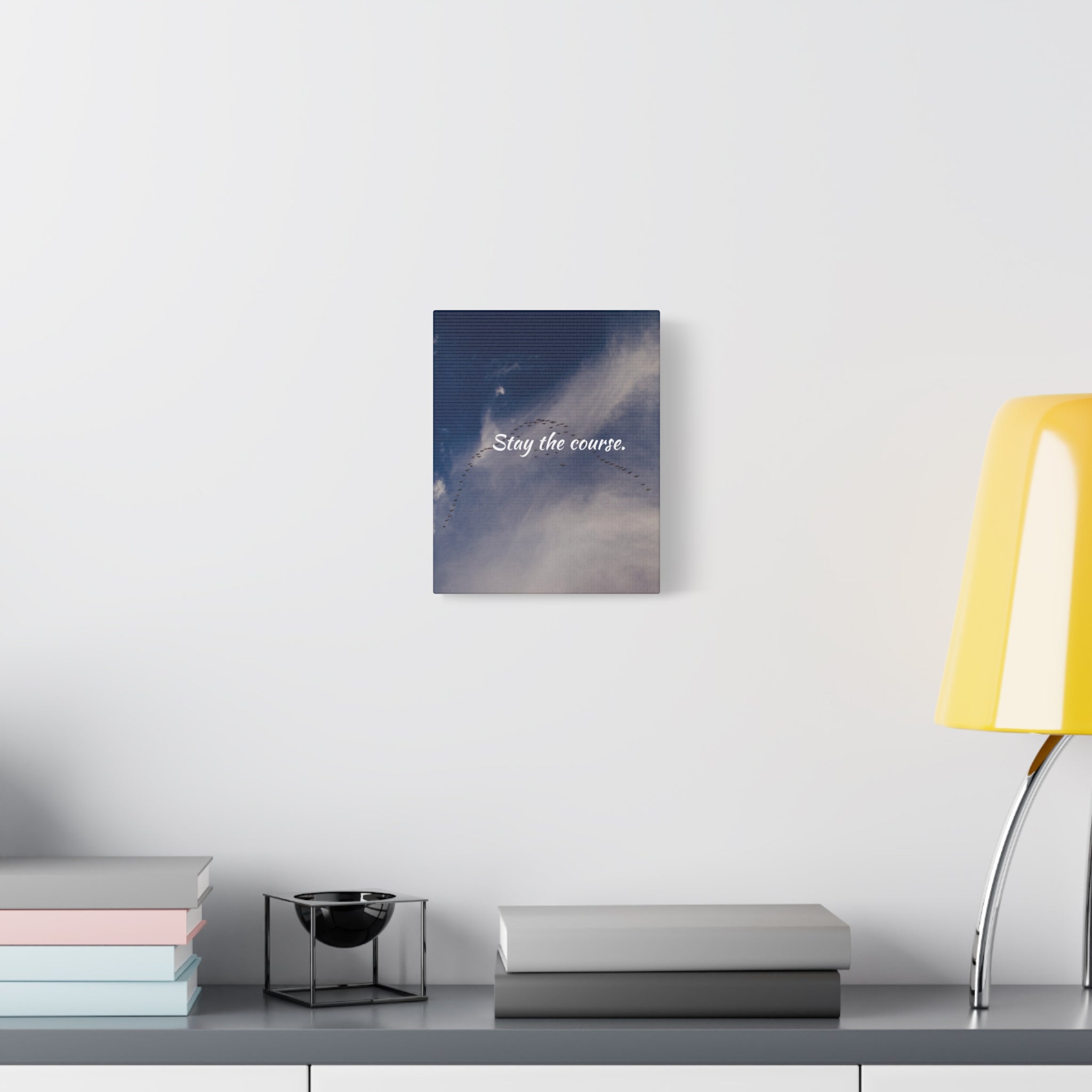 Canvas print with 'Stay the course' text on a wall above a desk with books and a lamp.
