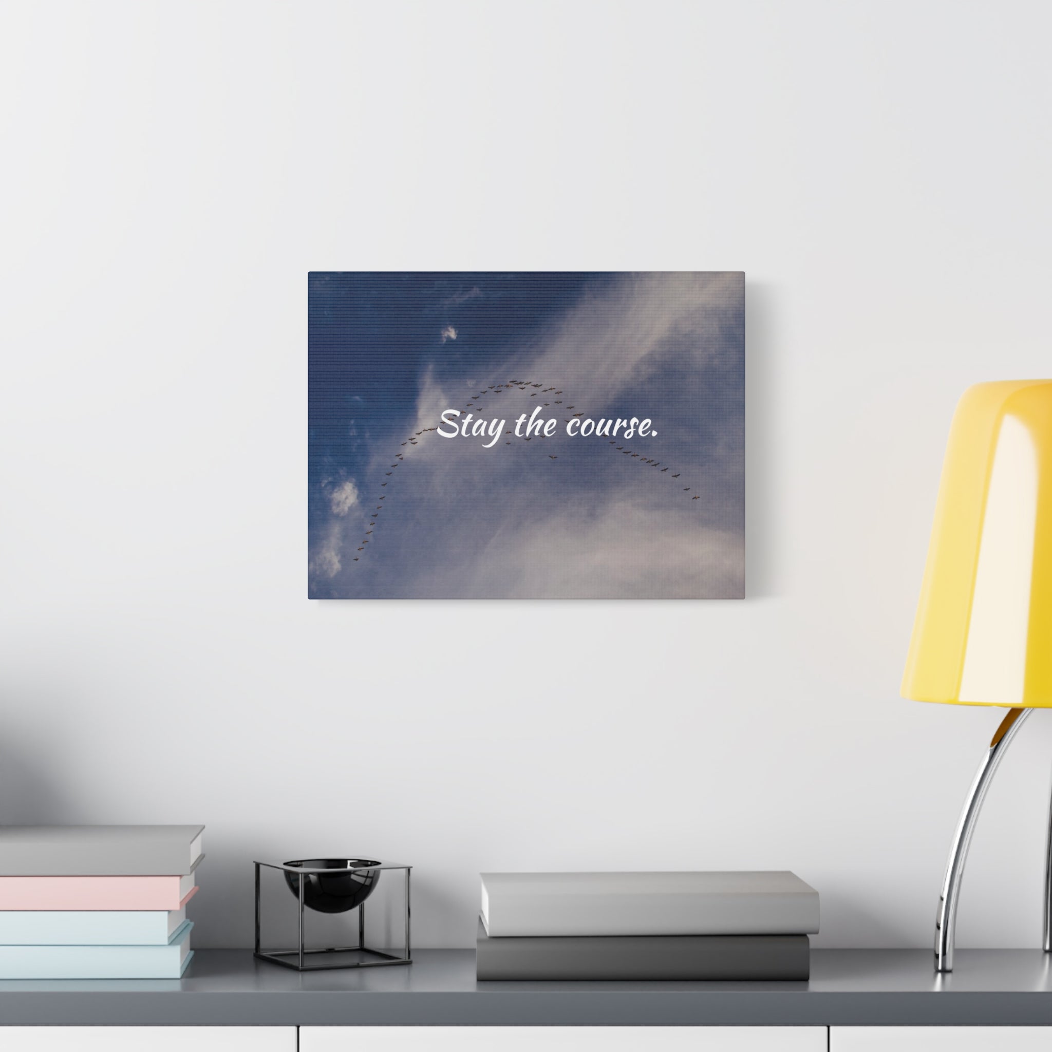 Canvas print with 'Stay the course.' text on a wall above a desk with books and a lamp.
