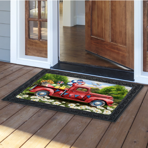 Decorative doormat with a red truck design on a wooden floor outside a house.