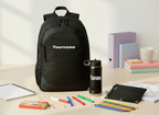 A black backpack and matching black water bottle, both printed with the customizable text 'Yourname', displayed on a light beige desk alongside school and office props including colored pencils, a notebook, binder folders, and a mug.