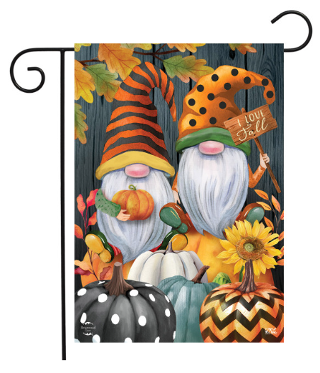 A decorative garden flag hanging from a black metal scroll-style stand. The flag features a fall-themed illustration with two gnome figures, pumpkins, sunflowers, autumn leaves, and the words 'I Love Fall' against a dark wood-plank background.