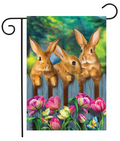 Decorative garden flag with three rabbits and flowers on a wooden fence.