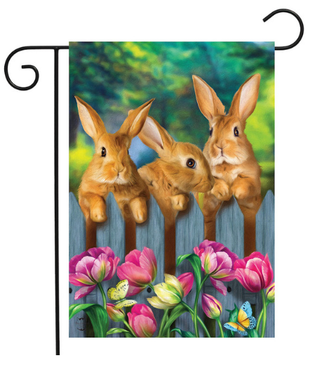 Decorative garden flag with three rabbits and flowers on a wooden fence.