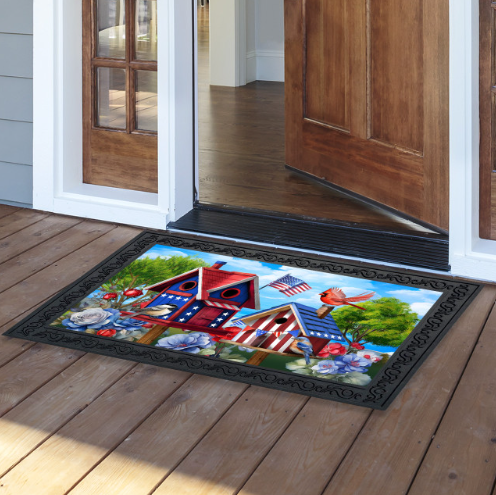 Decorative doormat with a colorful design of birdhouses, flowers, and an American flag on a wooden floor.