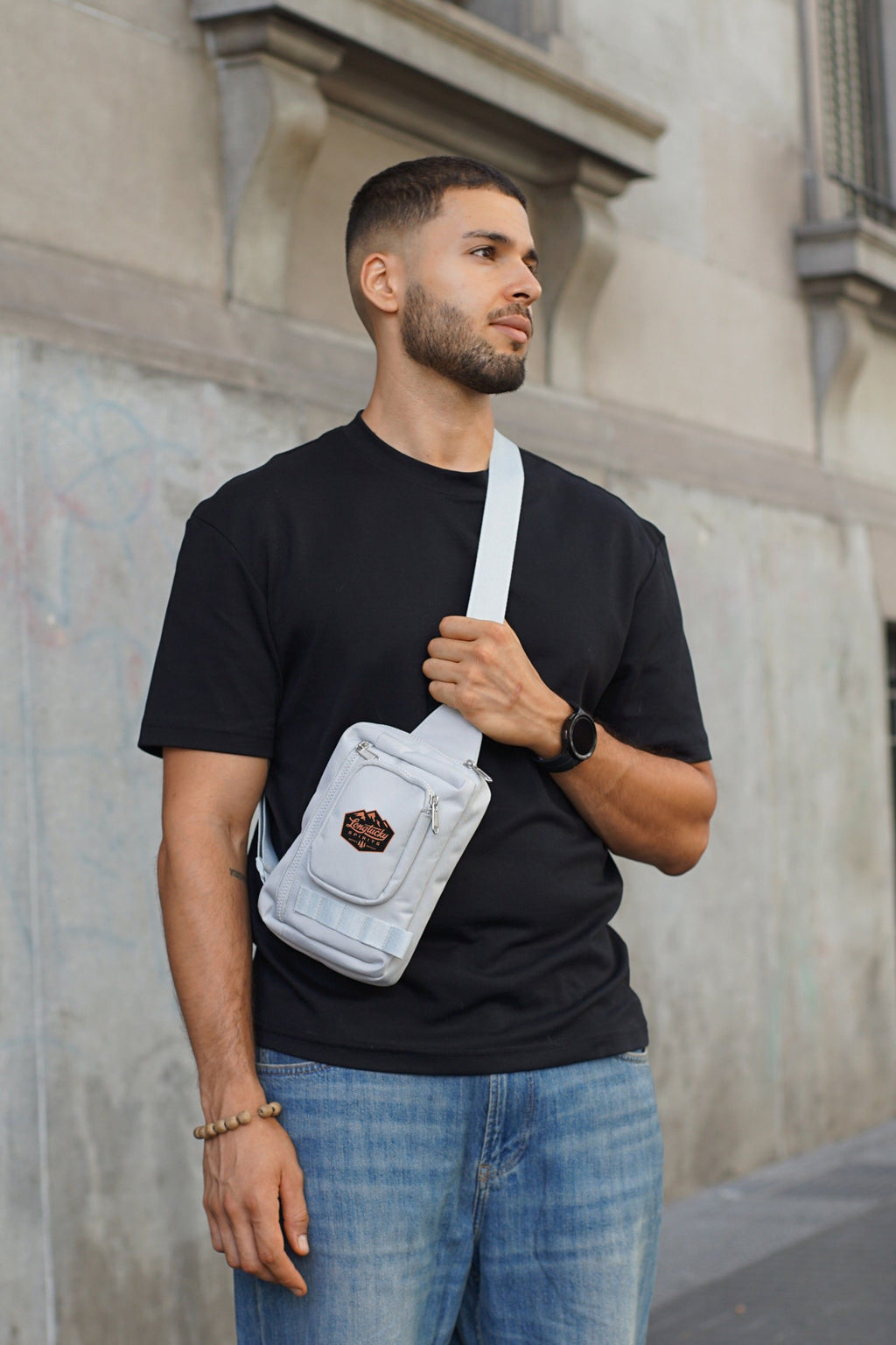 Man wearing a black t-shirt and blue jeans with a gray crossbody bag with design on front pocket against a neutral wall.