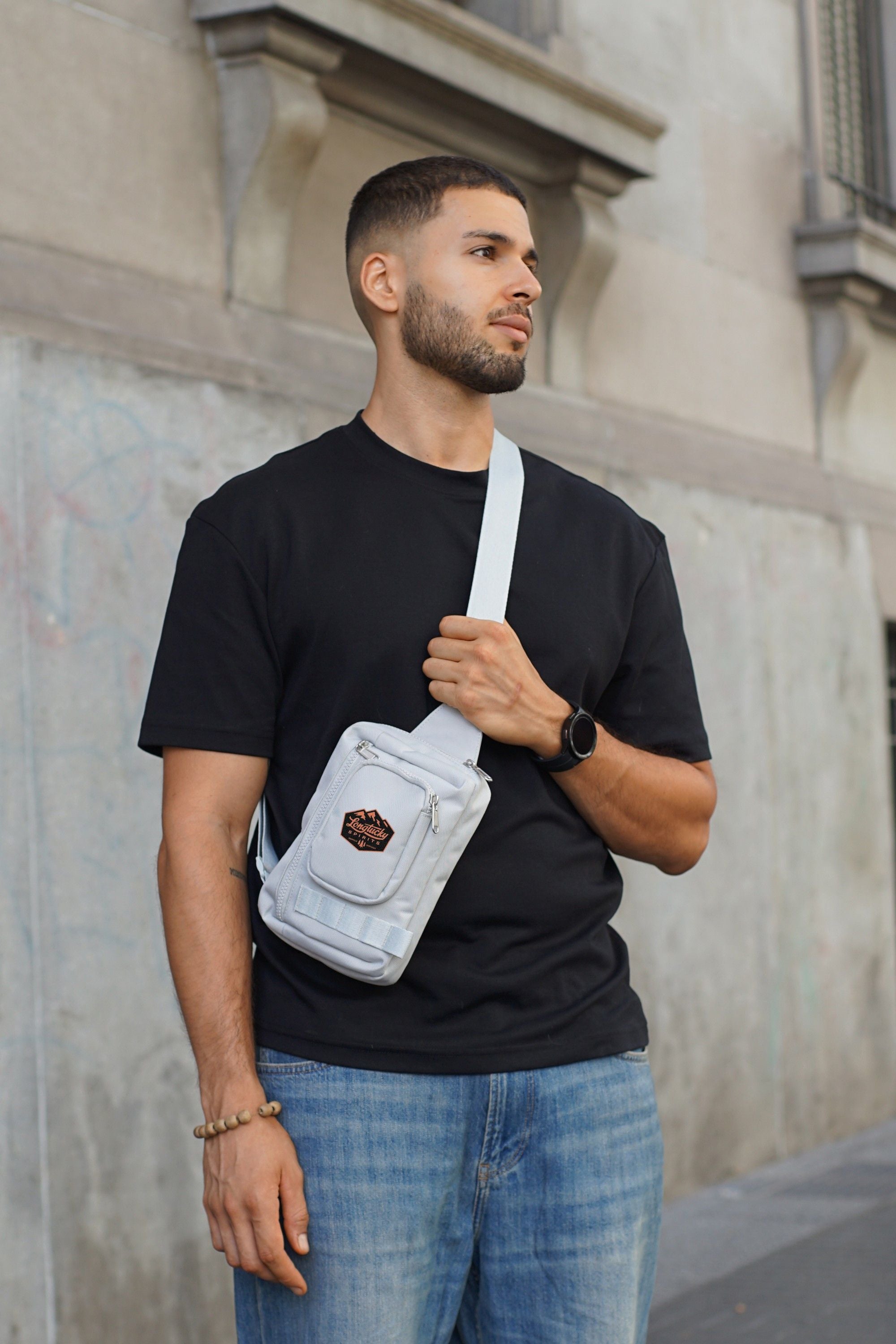 Man wearing a black t-shirt and blue jeans with a gray crossbody bag with design on front pocket against a neutral wall.