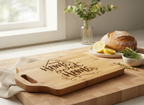 A rectangular wooden cutting board with a handle, engraved with the phrase 'Home is sweet Home' in a dark script, personalized with name, and styled on a larger wooden board in a bright kitchen setting with bread, lemons, herbs, and a vase of flowers.