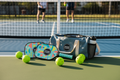 Pickleball equipment including bags and balls on a court with players in the background.