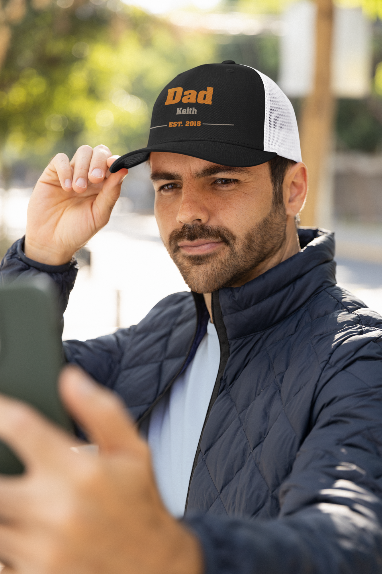 Man wearing a cap with 'Dad' embroidered on it, taking a selfie outdoors.