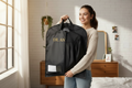 Woman holding a black garment bag with 'DR. AN' branding in a bedroom.