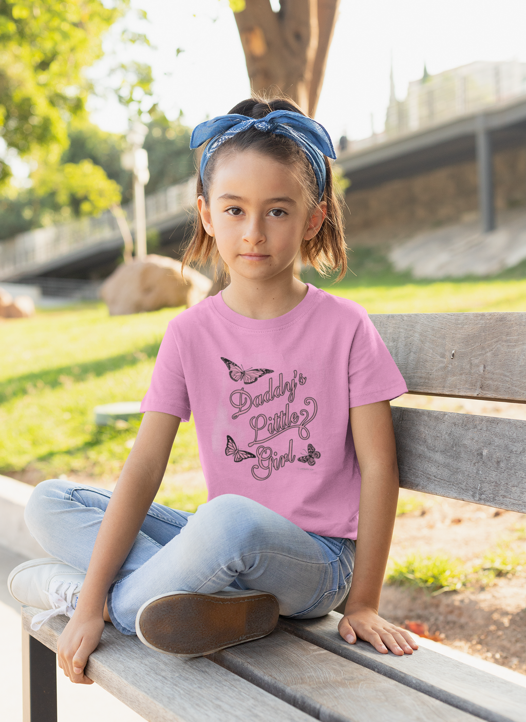 Young girl sitting on a park bench wearing a pink t-shirt with "Daddy's Little Girl" text and butterfly graphics.