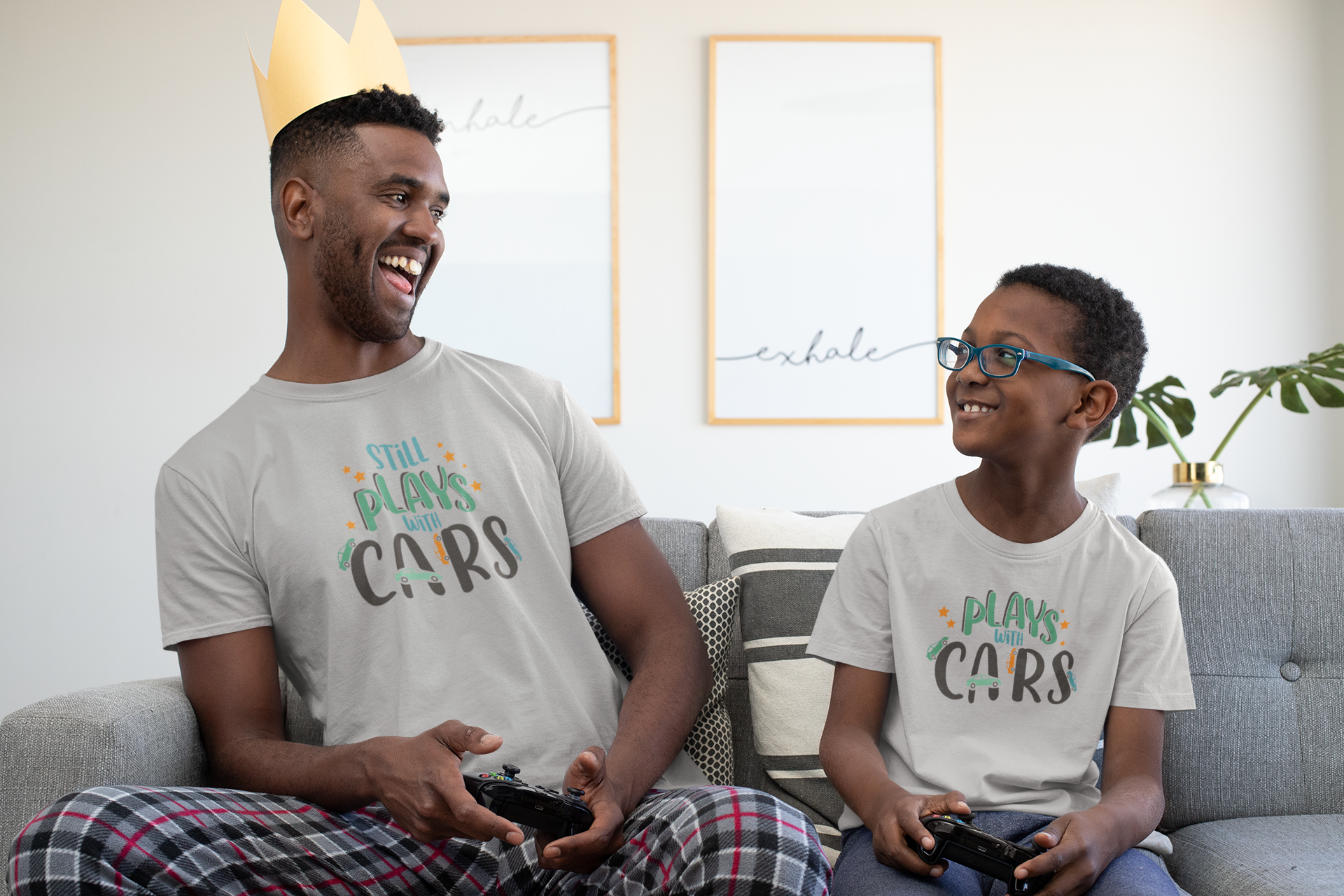 Father and son playing video games wearing matching shirts that say “Still Plays With Cars” and “Plays With Cars”