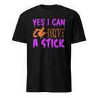 A black short-sleeve t-shirt with a graphic on the chest that reads 'YES I CAN DRIVE A STICK' in purple and orange text, featuring an orange silhouette of a witch riding a broomstick.