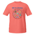 A coral pink short-sleeve t-shirt with a colorful graphic on the front that reads 'She is a teacher' surrounded by school-themed illustrations like books, bows, a globe, and flowers.