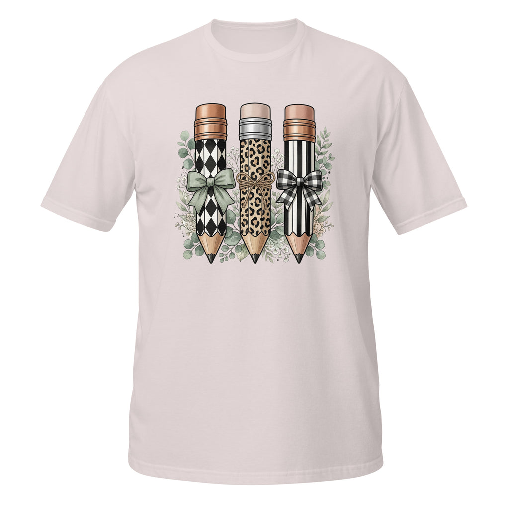 A light pink short-sleeve crewneck t-shirt featuring a graphic print of three decorative pencils with leopard, harlequin, and black-and-white striped patterns, accented with bows and green leaves.