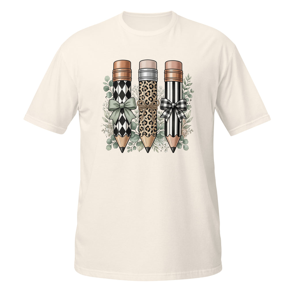 An off-white short-sleeve t-shirt with a graphic print of three decorative pencils (one with a black and white harlequin pattern, one with a leopard print, and one with a black and white striped pattern) adorned with bows and green leaves, displayed on a solid white background.