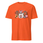 A bright orange short-sleeve crewneck t-shirt featuring a central graphic of a white dog wearing sunglasses and a pink polka-dot bow, surrounded by the word 'BOO' in leopard-print and plaid letters, with pumpkins, sunflowers, and hearts.