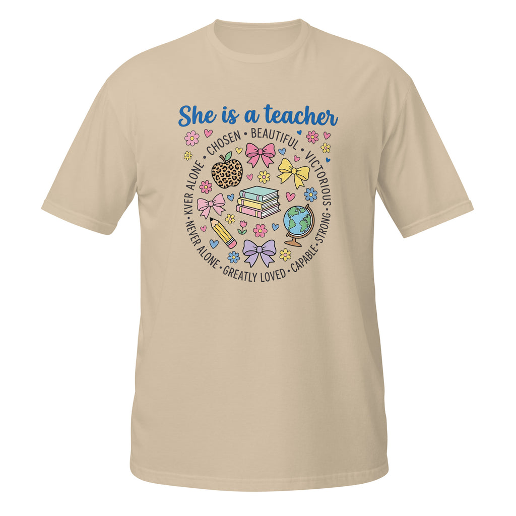 A sand short-sleeve t-shirt with a colorful graphic on the front that reads 'She is a teacher' surrounded by school-themed illustrations like books, bows, a globe, and hearts.