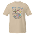 A sand short-sleeve t-shirt with a colorful graphic on the front that reads 'She is a teacher' surrounded by school-themed illustrations like books, bows, a globe, and hearts.