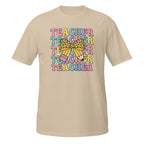 A beige short-sleeve crewneck t-shirt featuring a colorful graphic on the chest with the word 'TEACHER' repeated in pastel colors and a yellow leopard-print bow in the center.