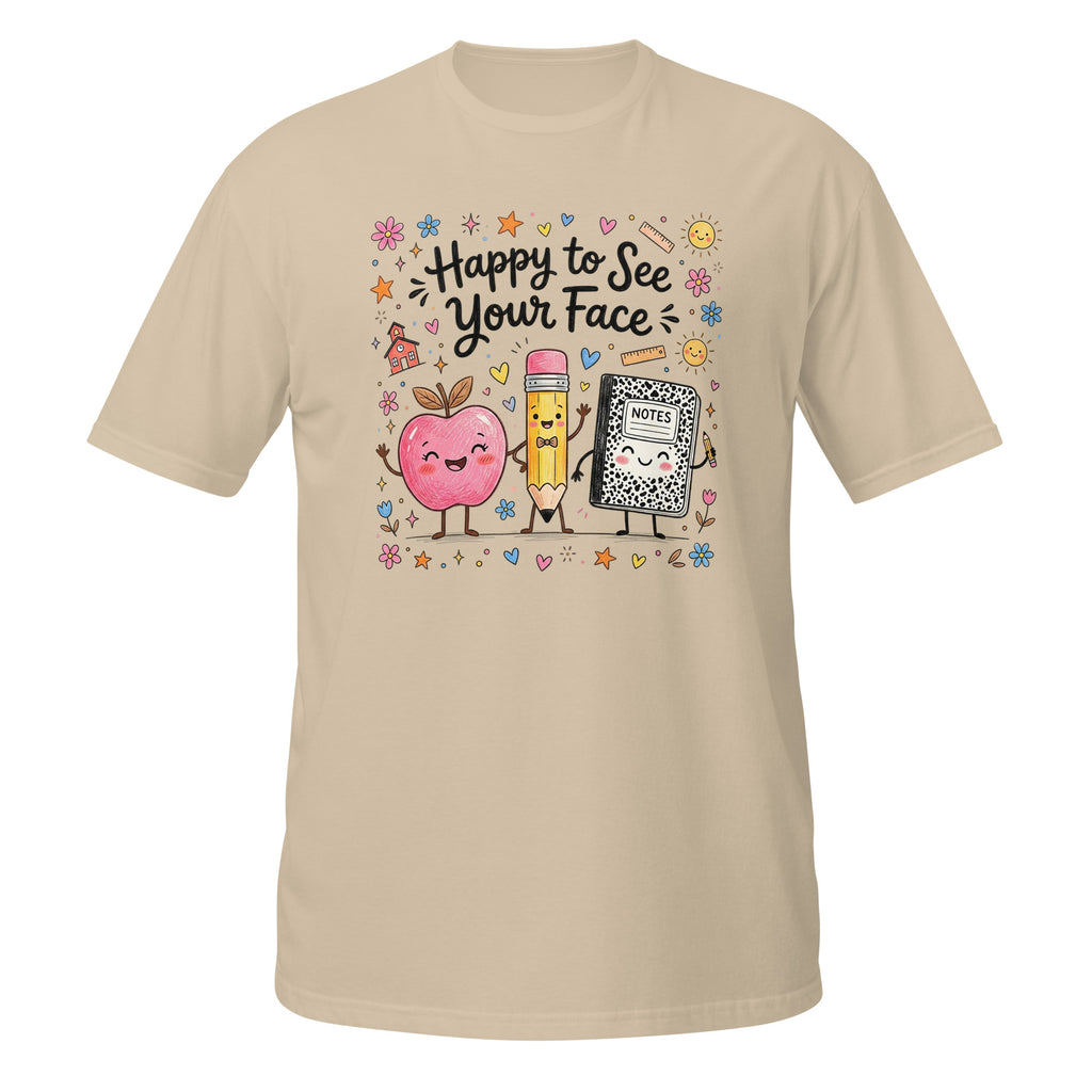 A beige short-sleeve crewneck t-shirt featuring a colorful cartoon graphic of a pink apple, a yellow pencil, and a black-and-white cat in the center, surrounded by the text 'Happy to See Your Face' and small decorative doodles.