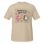 A beige short-sleeve crewneck t-shirt featuring a colorful cartoon graphic of a pink apple, a yellow pencil, and a black-and-white cat in the center, surrounded by the text 'Happy to See Your Face' and small decorative doodles.