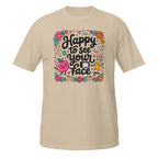 A beige short-sleeve t-shirt with a colorful graphic on the chest that reads 'Happy to see your face' in black script, surrounded by cute illustrations of a smiling pink apple, a yellow pencil, flowers, and school-themed doodles.