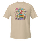 A beige short-sleeve crewneck t-shirt featuring a colorful graphic on the chest with the text 'Ready for Another Chapter' and an illustration of books, a smiling apple, and reading accessories.