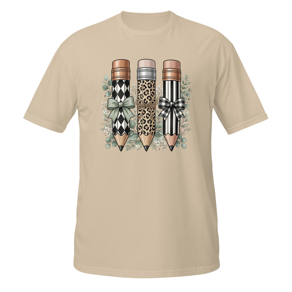A beige short-sleeve crewneck t-shirt featuring a graphic print of three decorative pencils with diamond, leopard, and striped patterns, accented with bows and green leaves.