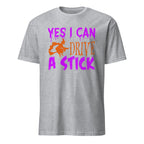 A sport grey short-sleeve t-shirt with a graphic on the front that reads 'YES I CAN DRIVE A STICK' in purple and orange text, featuring an orange silhouette of a witch riding a broomstick.