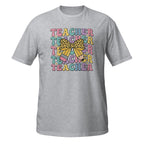 A heather gray short-sleeve crewneck t-shirt featuring a colorful graphic on the chest with the word 'TEACHER' repeated in pastel rainbow letters, overlaid with a yellow leopard-print bow and a pink pencil.